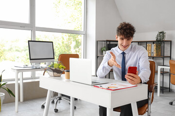 Young businessman with mobile phone video chatting at table in office