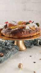 A festive King Cake with candied fruits and powdered sugar on a gold stand, surrounded by Christmas decorations