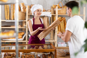 Hospitable smiling saleswoman in burgundy apron, working in cozy rustic bakery, engaging in friendly conversation over counter and offering fresh baguettes to male customer