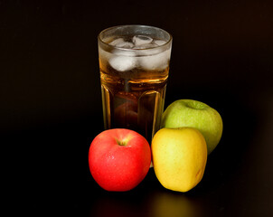 Apple juice in a tall faceted glass with ice on a black background, ripe fruits of different varieties nearby.
