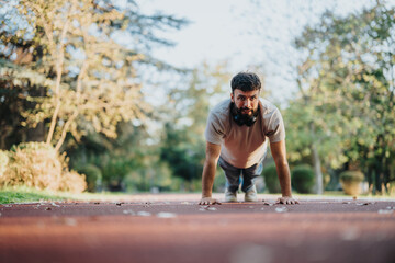 A determined man performs push-ups on a track in a sunlit park, surrounded by trees. This image...