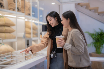 Two young female tourists with a camera and takeaway coffee cups are choosing coffee beans in a modern coffee shop, deciding which type of coffee they want to try