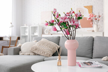 Vase with beautiful magnolia flowers and magazines on table near grey sofa in modern living room