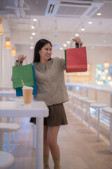 Happy customer showing colorful shopping bags while standing by a table in a modern food court, enjoying a relaxing break after a successful shopping spree