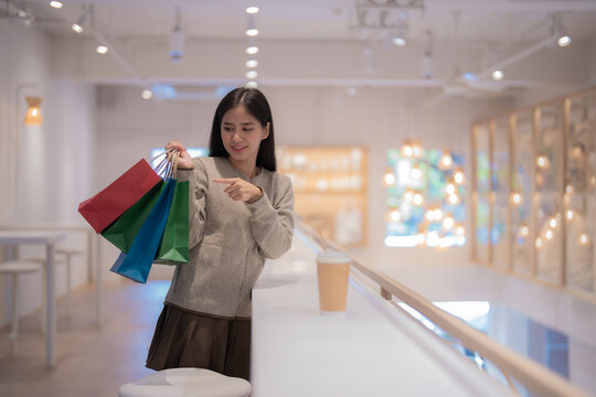 Young asian woman is pointing at her shopping bags while standing near a railing in a brightly lit mall, with a takeaway coffee cup resting on the surface beside her
