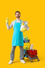 Male janitor with bucket and trolley of cleaning supplies on yellow background