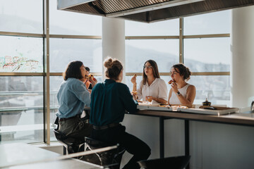 A group of colleagues sit at a kitchen counter in the office, enjoying a lunch break together. They...