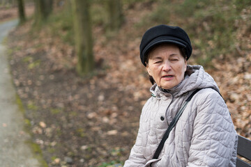 An elderly woman in a jacket and hat sits alone on a bench in a park. The concept of loneliness in old age