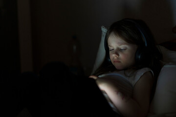 Little cute sad girl with headphones looking at tablet lying in bed in the dark. Concept of lonely modern digital life