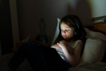 Sad little girl wearing headphones, lying in bed in the dark and looking at a tablet. Concept of...