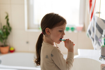 Cute little girl brushing her teeth with a toothbrush in the bathroom, standing by a large window. Concept of children’s health, hygiene, and daily care routines.