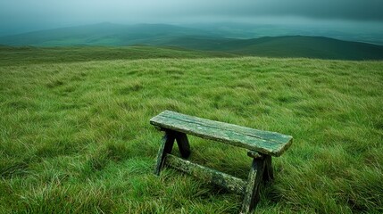 Fototapeta premium A solitary wooden bench rests on a lush green hillside, surrounded by a misty landscape and rolling hills under a cloudy sky.