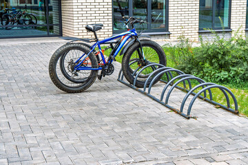 Mountain bike with thick tires parked in a metal bike rack on a paved area near a building, greenery in the background. Urban transport concept