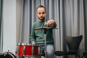 A drummer in a green sweater adjusts the cymbal in a practice studio, concentrating on his setup. The setting features a drum set and modern chair, evoking a sense of dedication to music artistry.