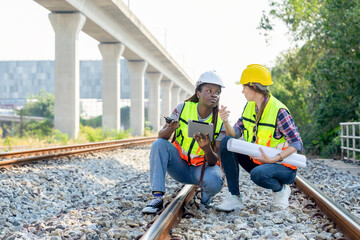 multi ethnic railway construction inspector working outdoors,at rail tracks,discussing work,african woman technician having walkie talkie and digital tablet,caucasian female holding blue print