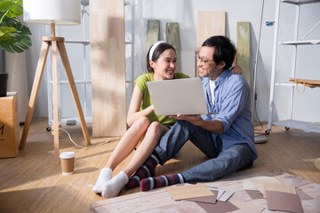 Asian couple sitting in home undergoing renovation, working together to choose furniture, wall colors, and decorations using a laptop. home, savings, loans, decoration, dreams concept.