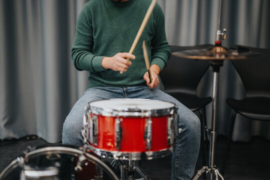 A musician wearing a green sweater is playing a red drum set. The focus is on the hands and drumsticks as they strike the drum in a studio environment.