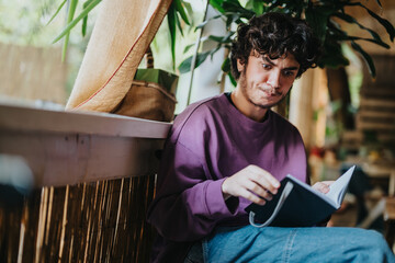 A young man sits indoors, deeply engrossed in a book. Sunlight filters through the windows, creating a warm and cozy atmosphere around him.