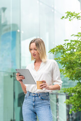 Fototapeta premium Caucasian Woman with Tablet While Having a Cup of Coffee In Front a Modern Glass Office Building