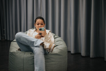 A young woman comfortably sits on a bean bag, engrossed in her smart phone. The setting conveys a...