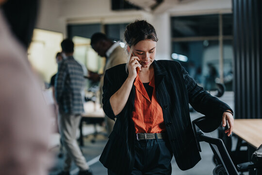 Confident businesswoman speaking on her mobile phone in a lively office setting, surrounded by colleagues. Image portrays a dynamic workplace atmosphere highlighting communication and collaboration. - Powered by Adobe