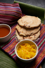 Pupusas with curtido and sauce served on a tamale leaf atop a wooden surface, highlighting traditional Central American flavors.