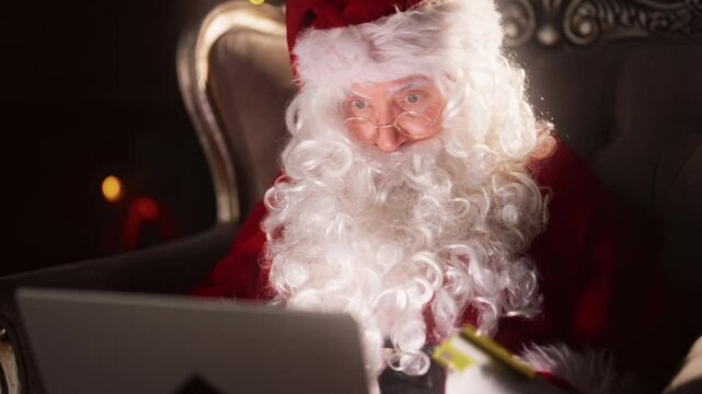 Funny old man in costume of Santa Claus ordering gifts, paying online, portrait . Online shopping in Christmas holiday, payment by credit card in web-store, face of wizard looking at computer display