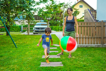 Happy child kicking inflatable ball with mother in garden during summer vacation