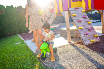 Happy child riding toy motorcycle with mother watching in backyard playground