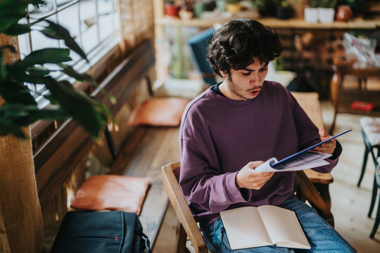 A young man with curly hair is intently reading a magazine while sitting in a warm and inviting coffee shop. The casual atmosphere suggests relaxation and focus amidst the cozy surroundings.