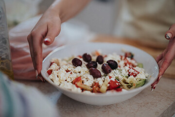 A delicious Greek salad with feta cheese, olives, and fresh vegetables is being prepared. Hands artistically arrange toppings in a kitchen setting, embodying Mediterranean cuisine and culinary