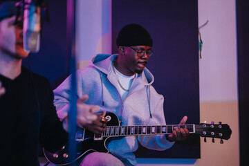 A young musician wearing a hoodie and beanie plays an electric guitar in a recording studio. The...