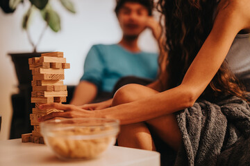 Two friends are engaged in a relaxed game night, playing Jenga with a bowl of snacks nearby. The cozy setting reflects leisure and companionship, capturing the essence of casual indoor gatherings.