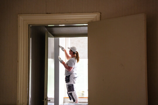 Female plasterer applying putty on wall seen through doorway