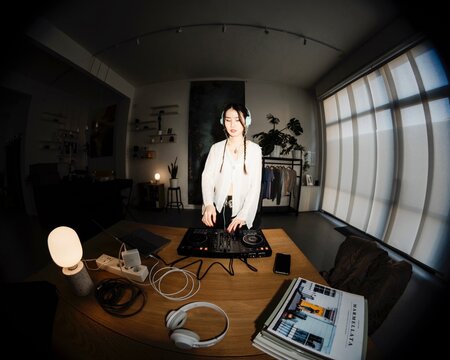 smiling young woman DJ wearing mint headphones mixes music on a DJ controller in a cozy home studio with shelves, houseplants, and decorative elements in the background.