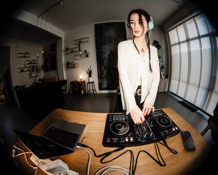 young woman DJ wearing mint headphones stands confidently at a DJ controller and laptop in a modern home studio with artistic decor, shelves, and indoor plants.