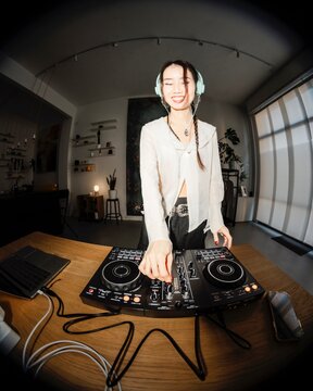 smiling young woman DJ wearing mint headphones mixes music on a DJ controller in a cozy home studio with shelves, houseplants, and decorative elements in the background.