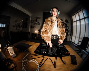 stylish DJ in a fur coat and teal headphones focuses on a professional DJ mixer placed on a desk, surrounded by cables, a laptop, and warm ambient lighting.