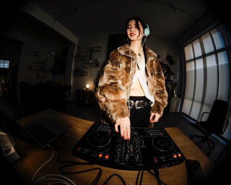 stylish DJ in a fur coat and teal headphones focuses on a professional DJ mixer placed on a desk, surrounded by cables, a laptop, and warm ambient lighting.