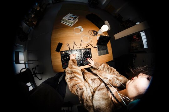 Overhead fisheye view of a DJ in a fur jacket using a professional mixer setup on a wooden table, surrounded by tech devices like a laptop, cables, and books.