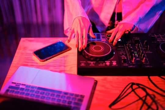 DJ&rsquo;s hands operate a professional mixer on a wooden table, with a smartphone and laptop nearby, under vibrant pink and blue lighting in a club performance setting.
