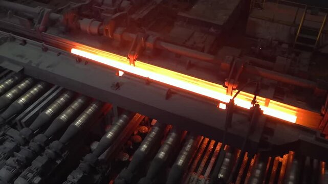 Witness the intense process of hot steel billets being transported on a conveyor in a steel mill. The glowing metal highlights the industrial operation.