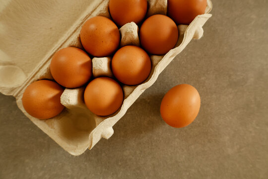A top view of brown eggs neatly arranged in a carton with one egg placed outside on a kitchen countertop. The warm lighting highlights the smooth texture of the eggshells.. - Powered by Adobe