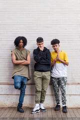 Obraz premium full body of multiethnic teenager friends with folded hands leaning against white brick wall, concept of friendship in youth and urban lifestyle, copyspace for text