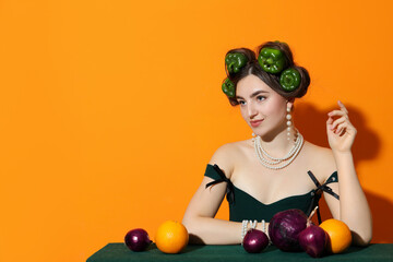 Young woman with peppers in her hair, fruits and vegetables at green table on orange background,...