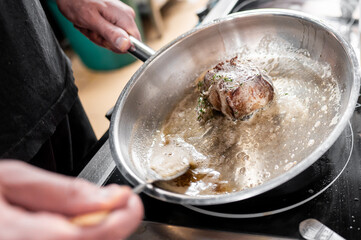 A close-up of a chef cooking a piece of meat in a stainless steel pan. The meat is sizzling in hot butter, with herbs adding flavor, creating an appetizing and inviting atmosphere.