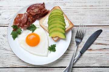 Romantic breakfast with fried bacon, heart shaped egg and avocado toast on white wooden table, flat lay. Valentine's day celebration