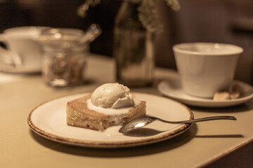 Piece of cake topped with a scoop of ice cream on a table in a blurry cafe or restaurant setting. a place to enjoy a leisurely meal or a cup of coffee