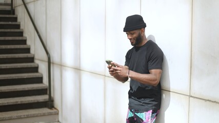 Smiling young man using smartphone leaning against wall next to stairs