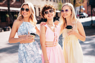 Three young beautiful smiling hipster female in trendy summer stylish clothes. Sexy carefree women posing in the street. Positive models having fun, hugging, in sunny day, drink tea, coffee